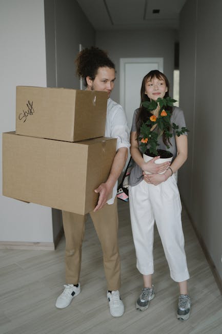 A woman and a young girl are standing inside a narrow hallway during a home relocation process. The woman is carrying two large cardboard boxes, one stacked on top of the other, and appears to be prep