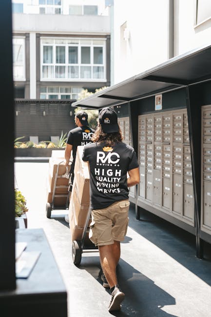 A woman and a young girl are standing inside a narrow hallway during a home relocation process. The woman is carrying two large cardboard boxes, one stacked on top of the other, and appears to be prep