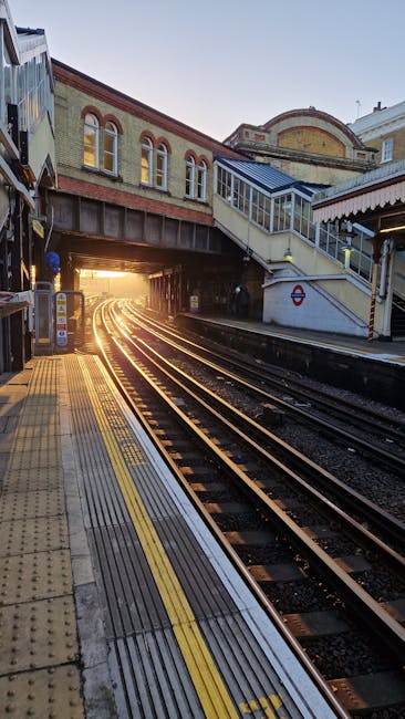 A photograph of an underground London tube station platform at Goodge Street, showing beige tiled walls and a curved ceiling with metal panels. To the left, there is a staircase with black metal raili